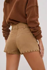 Short de efecto gamuza color camel, de tiro medio, con detalle de tachas metálicas doradas envejecidas en los bolsillos y en el ruedo. Incluye cinturón a tono con hebilla metálica.