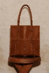 Bolso tipo tote de cuero vacuno color suela, confeccionado artesanalmente con costuras decorativas visibles en los bordes y en el centro. Cuenta con doble asa larga para llevar al hombro y detalles de tiras colgantes en la unión de las asas.