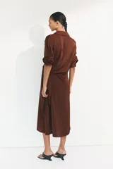 Vestido midi camisero de sarga liviana con brillo satinado, color marrón, de manga larga, con hombros caídos, botonadura frontal y puños sencillos. Presenta paneles drapeados cruzados en la cintura con tiras para atar al costado.