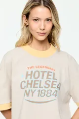 Remera de corte amplio, color beige, con mangas cortas y cuello redondo. Presenta un estampado frontal con la inscripción "The Legendary Hotel Chelsea NY 1884" en tonos celeste y marrón, con detalles en amarillo en el cuello y los puños.
