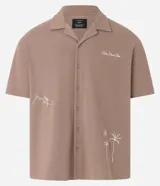 Camisa de manga corta, color marrón topo, con cuello camisero y corte amplio (boxy). Presenta bordados minimalistas en blanco en el frente, simulando un paisaje.
