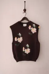 Chaleco de punto de lana merino color marrón oscuro, con diseño sin mangas, cuello redondo y bordados artesanales de flores blancas y rosas con tallos verdes.
