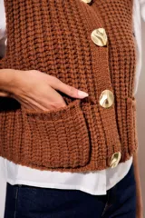 Chaleco tejido color beige con cuello redondo, sin mangas, con cierre frontal de cuatro botones dorados y dos bolsillos frontales.