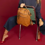 Mochila de cuero sintético con textura granulada, color verde oliva. Cuenta con dos bolsillos frontales con cierre metálico, asa superior rígida y correas ajustables de cinta textil. Incluye placa metálica decorativa en la parte inferior.