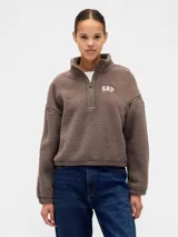 Buzo de polar tipo sherpa color marrón, con cuello alto y cierre frontal parcial. Presenta el logo de la marca bordado en el pecho y detalles de costuras reforzadas en hombros y mangas.