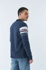 Remera tipo polo de manga larga azul marino con rayas horizontales blancas, grises y rosadas en el pecho. Tiene cuello clásico con botones y logo estampado en el pecho. Corte regular.