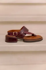 Sandalia de cuero bordó con diseño tipo thong, tiras anchas cruzadas con tachas metálicas doradas, tira con hebilla ajustable en el talón y taco bajo cuadrado con acabado artesanal.