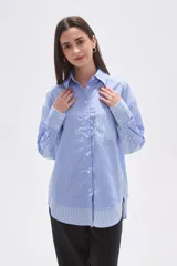 Camisa oversize de poliéster, con cuello camisero y cierre frontal de botones. Presenta un diseño de rayas verticales azules y blancas, con bloques de rayas horizontales en los puños y el ruedo inferior.