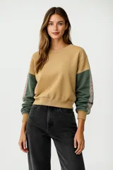 Buzo de tejido de punto color camel, con cuello redondo y mangas largas. Las mangas son de color verde oscuro y presentan una cinta decorativa con patrón étnico en tonos rosados y verdes.