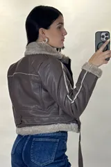 Campera tipo aviador de cuero sintético negro con interior y detalles de piel sintética al tono en cuello, puños y ruedo. Presenta cierre frontal cruzado y cinturón ajustable con hebilla metálica en la cintura.