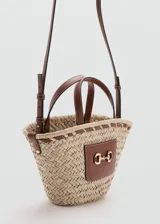 Bolso tipo cesto de fibra natural trenzada color beige, con detalles de cuero sintético marrón, doble asa corta de mano y correa bandolera ajustable y desmontable. Incluye un detalle de herraje metálico dorado en el frente sobre un bolsillo de cuero sintético.