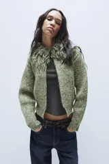 Chaqueta de punto de textura jaspeada en color verde, con cuello redondo adornado con flecos del mismo tejido, manga larga y cierre frontal con corchete metálico.