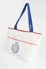 Bolso tipo shopper color blanco con detalles en rojo, confeccionado en algodón. Presenta un estampado central en azul con el escudo nacional y la frase 'El País de los mates y las siestas'. Cuenta con correas de hombro en color azul.