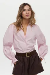 Camisa blanca de manga larga abullonada con detalle de punto smock en los puños y cordones para atar. Presenta cuello clásico y cierre frontal de botones.