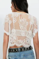 Top corto blanco confeccionado en encaje de nailon con diseño floral transparente, escote redondo y mangas cortas holgadas con terminación festoneada.