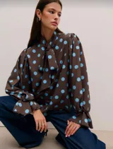 Camisa de manga larga con estampado de lunares celestes sobre fondo marrón. Presenta cuello con lazo para anudar, mangas amplias con puños acampanados y cierre frontal con botones.