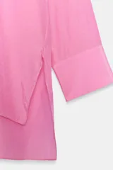 Blusa fluida de color rosado con cuello redondo ajustable mediante lazadas del mismo tejido, manga larga con puño y bajo asimétrico con aberturas laterales.