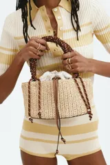 Cartera tipo tote de paja trenzada, con estructura rígida y base redonda. Cuenta con una bolsa interior de lona color crudo con cierre ajustable mediante cordones de cuero. Posee una manija superior de cuerda marrón trenzada y detalles metálicos dorados.