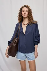 Camisa de manga larga azul marino con cuello clásico, botonadura frontal, mangas con sutil volumen y puños elásticos. Su silueta es amplia y de caída fluida.