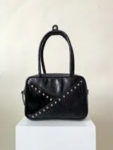 Cartera rígida negra con detalles en rojo y tachas plateadas. Tiene herrajes y cierre metálico plateado marca YKK, asa fina para llevar al hombro o en la mano y correa larga extra regulable. El interior es forrado, tiene un bolsillo con cierre enmarcado en cuero, y un segundo bolsillo plaqué.