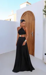 Vestido largo negro de corte sirena con diseño cut-out en la cintura, tirantes cruzados tipo halter al cuello y detalle de hebilla dorada central.
