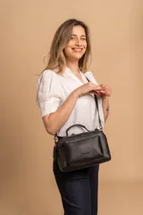 Bandolera de diseño rectangular en color negro, confeccionada en material sintético con textura. Cuenta con un asa superior fija, correa larga ajustable y extraíble, y dos bolsillos frontales, uno de ellos con cierre horizontal. Incluye el logo de la marca grabado en el frente.