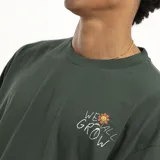 Remera marrón con estampado pequeño en el pecho que dice "We all grow" con una flor.