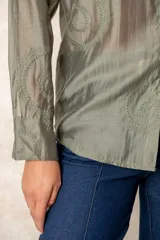 Camisa de manga larga en color blanco, confeccionada en tejido traslúcido con delicados bordados florales en hilo de seda al tono. Presenta cuello clásico, cierre frontal con botones y puños abotonados.