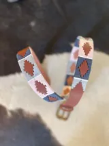 Cinturón de cuero en tono tostado con bordado a mano de hilos encerados en tonos marrones y beige con motivos geométricos, y hebilla metálica rectangular.