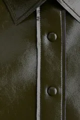 Camisa de manga larga color verde oscuro con acabado brillante (efecto charol o vinilo), cuello clásico y botones forrados al frente. Presenta costuras visibles que definen paneles y un bajo redondeado.