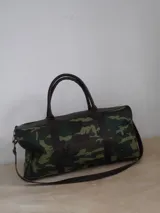 Bolso de viaje tipo duffel de lona negra con detalles en cuero tostado, asas de mano y correa ajustable para el hombro.