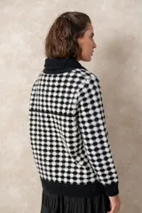 Chaqueta de punto grueso con patrón geométrico de cuadros en blanco y negro. Presenta cuello camisero, cierre frontal con cremallera y tirador circular, bolsillos frontales y puños acanalados.