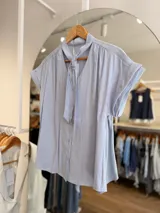 Blusa celeste con lazo en el cuello, escote en V, cierre frontal con botones y mangas cortas con vuelta.