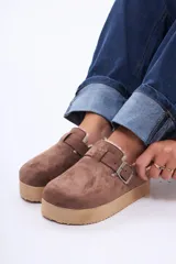 Sueco de gamuza color marrón con interior forrado en piel sintética. Presenta una tira ajustable con hebilla metálica en el empeine y una suela de plataforma acanalada en tono beige.