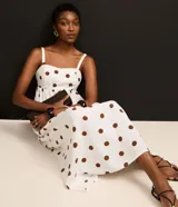 Vestido midi evasé sin mangas, confeccionado en tejido airflow color blanco con estampado de lunares marrones. Presenta escote corazón, breteles anchos con elásticos y lastex en la espalda.