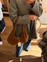 Cartera tipo hobo de textura gamuzada en color marrón oscuro, con diseño flexible y asa larga regulable. Presenta un detalle de flecos largos desmontables en uno de los laterales y cierre superior.