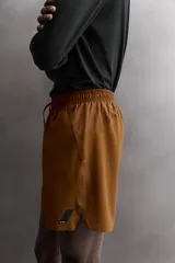 Pantalón corto deportivo color marrón oliva, confeccionado en tejido técnico ligero y elástico, con forro interior. Presenta cintura elástica ajustable con cordón, bolsillos frontales, bolsillo de seguridad trasero con cierre y aberturas laterales en el bajo.