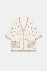 Blusa de escote en pico y manga corta, confeccionada en tejido color crudo con detalles de bordados florales y geométricos a contraste. Presenta cierre frontal con botones y bolsillos de plastrón en el delantero.