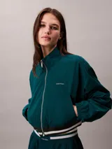 Campera tipo track Calvin Klein de tejido plano en color verde oscuro, de corte cropped y manga larga. Cuenta con cuello alto, cierre frontal de cremallera metálica, bolsillos laterales y elástico acanalado a rayas en los puños y el dobladillo. Presenta el logo de la marca bordado en el pecho.