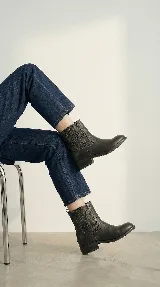 Bota de caña alta color negro, con diseño versátil que incluye un arnés desmontable con hebilla y argollas metálicas. Presenta costuras decorativas en la parte superior de la caña y un taco bajo cuadrado.