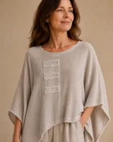 Blusa beige de corte holgado y asimétrico, con mangas amplias y tres parches rectangulares decorativos en el pecho. Presenta detalles de flecos en el cuello, dobladillo y mangas.