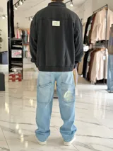 Buzo gris oscuro de corte oversize con cuello alto y cierre frontal parcial. Cuenta con mangas largas y puños elásticos, además de un pequeño bordado con texto en el pecho.