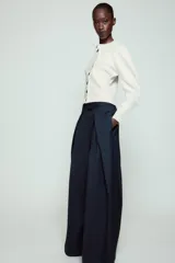 Pantalón de tiro medio de corte wide leg, confeccionado en mezcla de lyocell con patrón de rayas diplomáticas. Presenta bolsillos laterales ocultos, pliegues delanteros marcados y cierre frontal con cremallera, botón y ganchos metálicos.