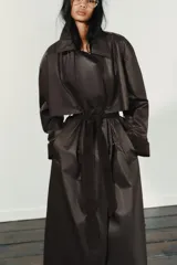 Gabardina larga de efecto encerado en color marrón oscuro, con cuello solapa y cierre frontal cruzado con botones. Presenta mangas largas con puños vueltos en pana, bolsillos laterales y cinturón ajustable de pana. Incluye pinza en la espalda.