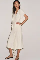 Falda midi de tiro medio, color blanco off-white, confeccionada en hilatura de viscosa. Presenta un diseño fluido, con bajo asimétrico y detalle de caída drapeada en el lateral. Cierre con cremallera oculta en costura.
