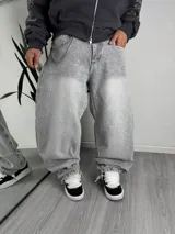 Pantalón jeans estilo baggy de corte muy holgado y tiro medio, con efecto acid wash en tono gris deslavado. Cuenta con una cadena metálica decorativa colgada de la presilla y botamangas con elástico fruncido.