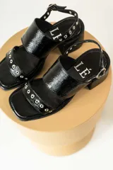 Sandalias de gamuza color marrón con taco cuadrado de madera. Presentan dos tiras anchas, una superior lisa y una inferior decorada con ojales metálicos plateados. Cuentan con cierre de pulsera con hebilla ajustable en el tobillo.