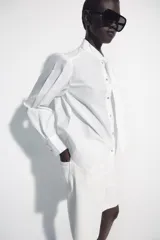 Camisa de algodón color blanco, con cuello subido y detalle de pañuelo incorporado, manga larga con puños anchos y cierre frontal con botones joya dorados.