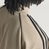 Campera deportiva Adidas SST TT color beige, con cuello alto, cierre frontal y las clásicas tres tiras negras en las mangas. Incluye el logo de Adidas bordado en el pecho.