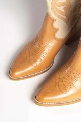 Bota texana de cuero color marrón claro con detalles en beige. Diseño con puntera en punta y caña media con bordados decorativos. Taco bajo de madera.